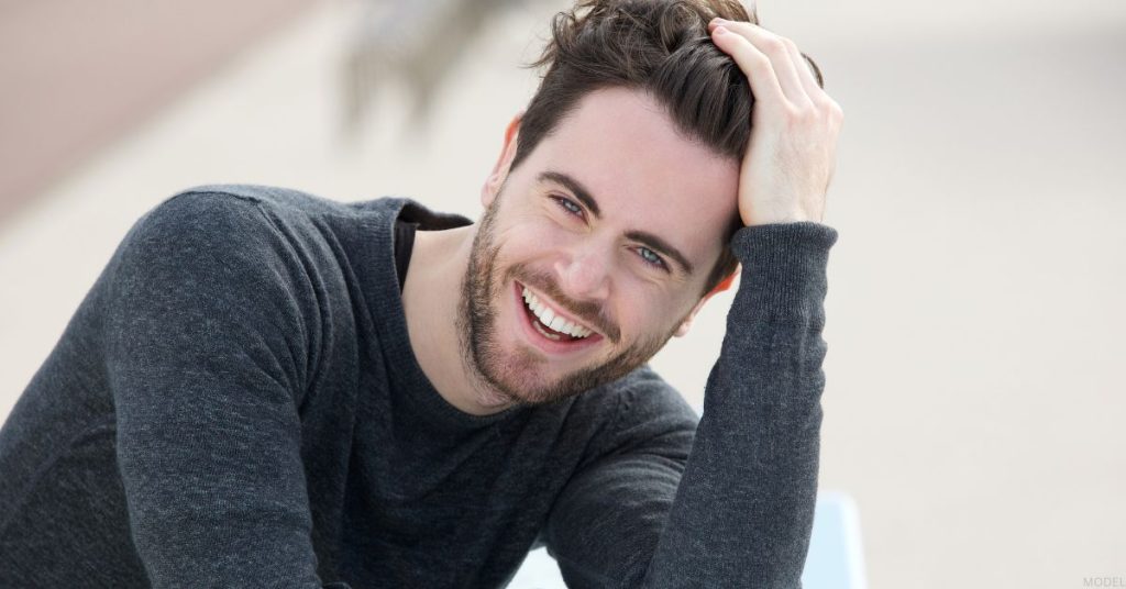 Man with brown hair smiling as he gently touches his hair. (MODEL)